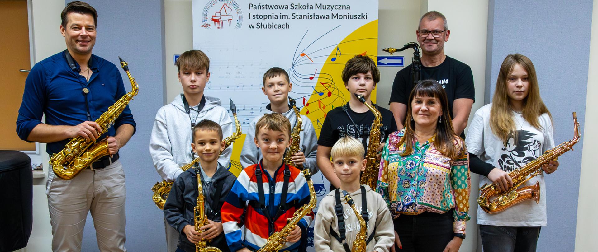 Uczestnicy warsztatów stoją w auli szkolnej wraz z prowadzącym, nauczycielem klasy saksofonu i Dyrektorką szkoły.