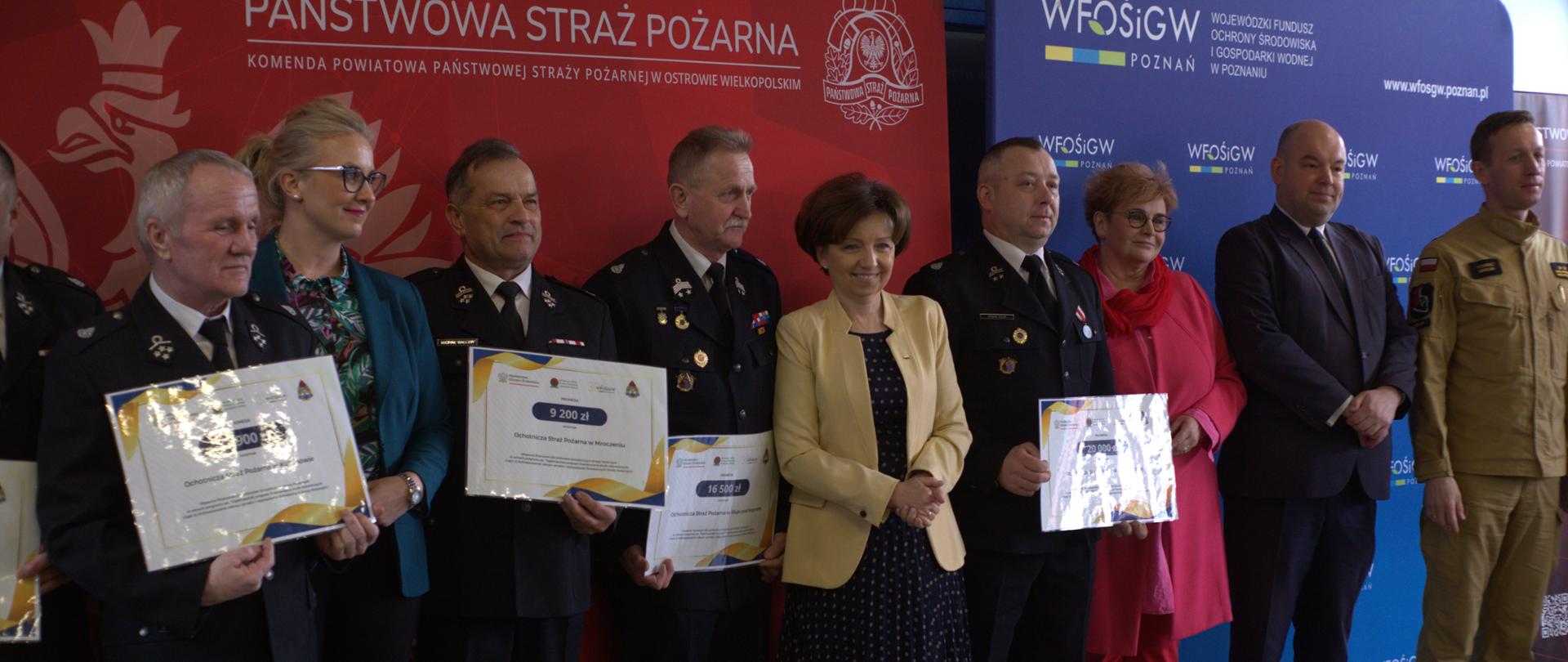 Zdjęcie przedstawia parlamentarzystów i strażaków podczas wręczania promes. Osoby stoją na tle banerów WFOŚiGW w Poznaniu i KP PSP w Ostrowie Wielkopolskim.
