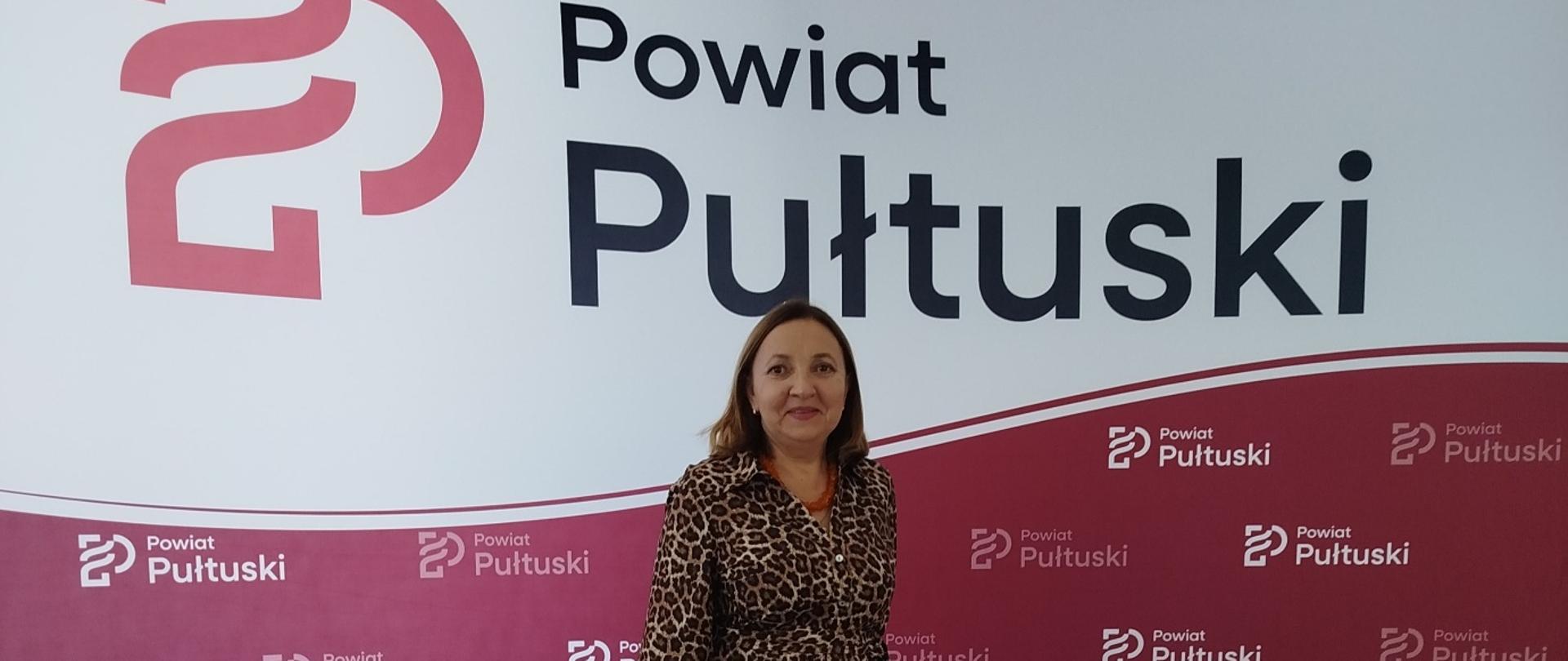 Kobieta w sukience w panterkę stoi przed ścianką promocyjną z logo i napisem „Powiat Pułtuski”.