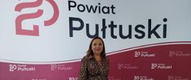 Kobieta w sukience w panterkę stoi przed ścianką promocyjną z logo i napisem „Powiat Pułtuski”.