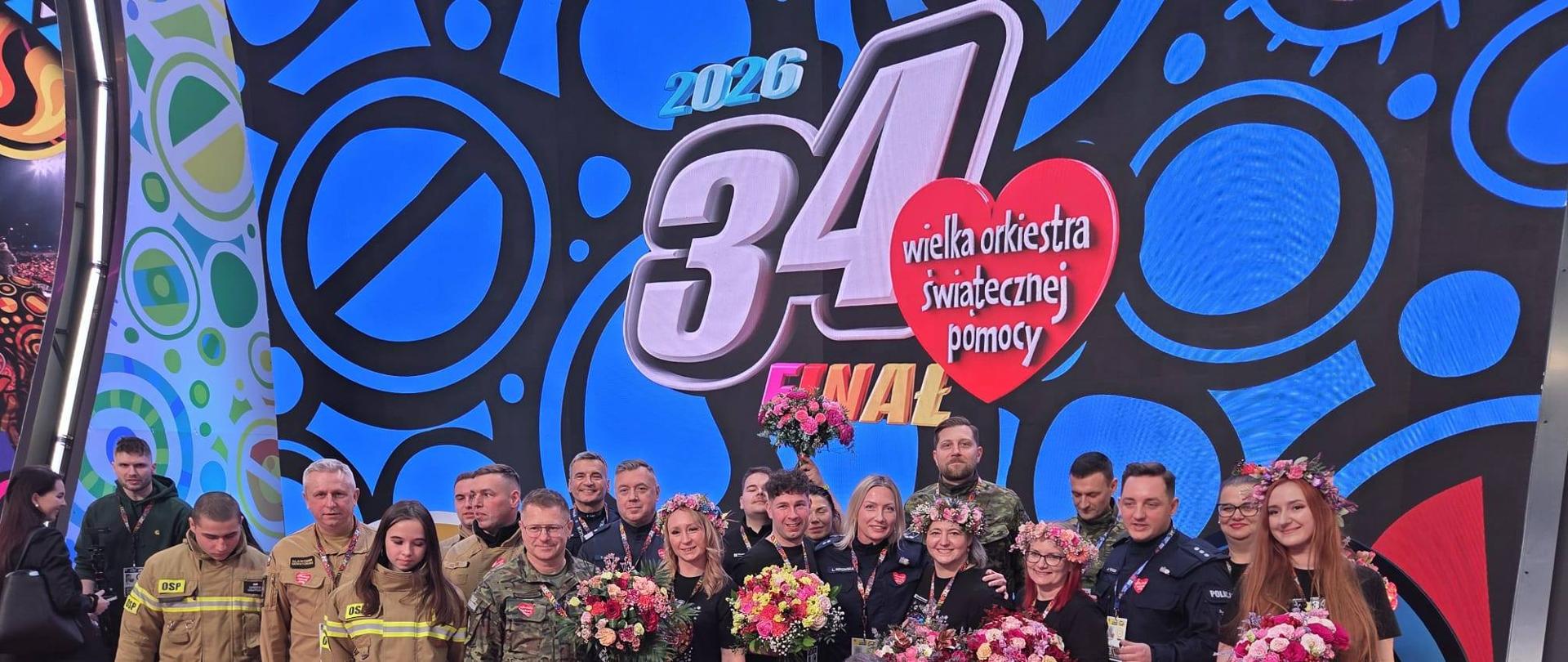 Grupowe zdjęcie wolontariuszy, strażaków, policjantów i żołnierzy na scenie finału 34. Wielkiej Orkiestry Świątecznej Pomocy, osoby trzymają kolorowe bukiety kwiatów.