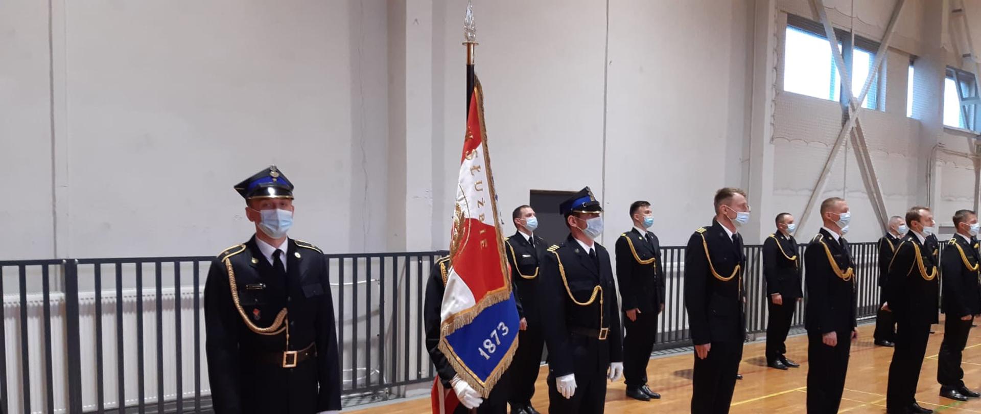 Zdjęcie przedstawia strażaków ubranych w mundury wyjściowe, jeden z nich trzyma sztandar. Zdjęcie wykonane na sali gimnastycznej.