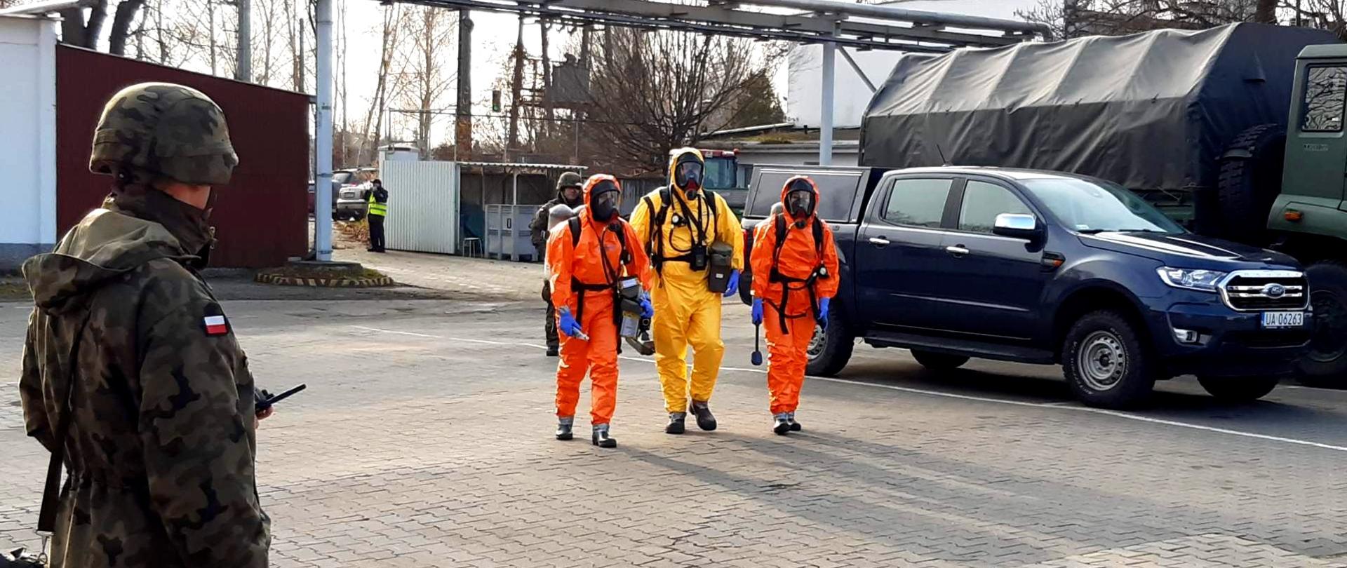 Trzech strażaków w kombinezonach ratownictwa chemicznego idzie przez plac, po lewej stronie stoi żołnierz, po prawo widoczne dwa samochody, w tle blaszane wiaty