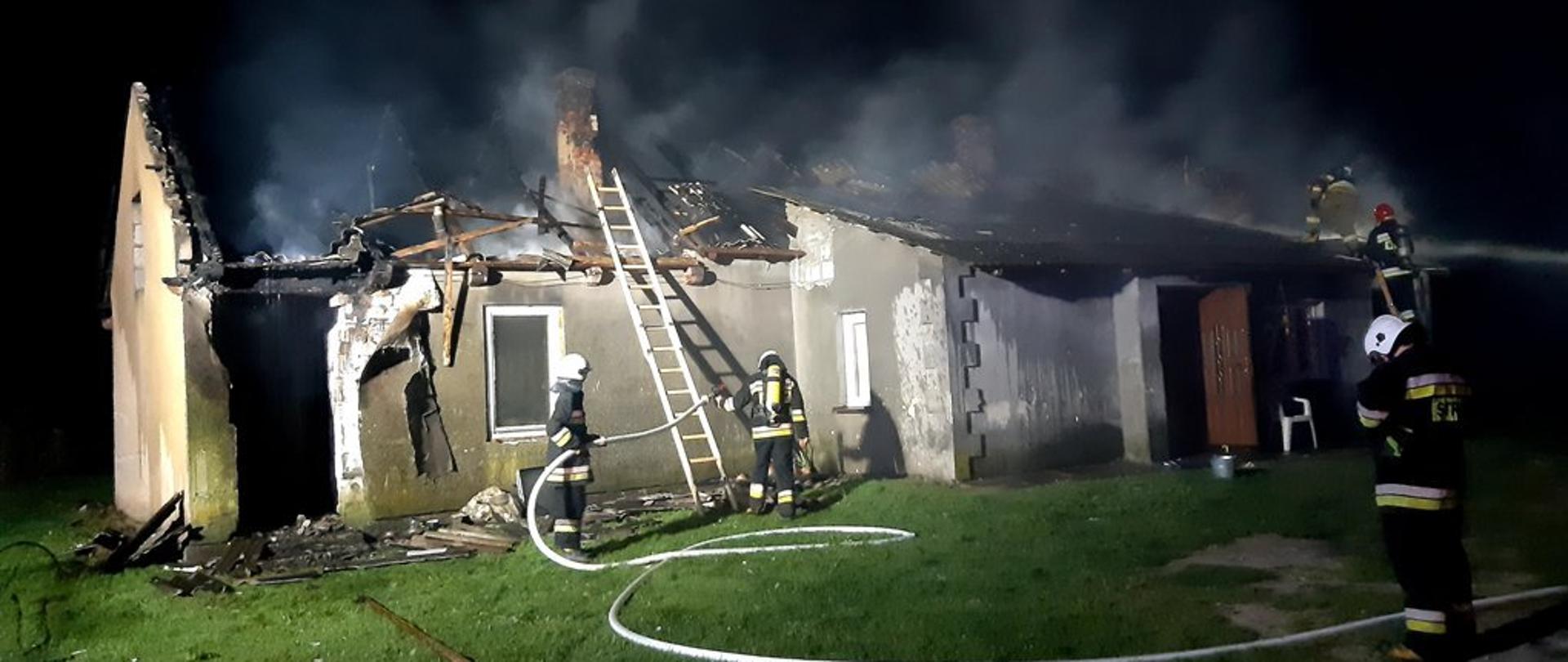 Na zdjęciu budynek mieszkalny ze spaloną więźbą dachową, o budynek oparta drabina strażacka oraz rozwinięta linia gaśnicza,przy budynku strażacy prowadzący działania gaśnicze