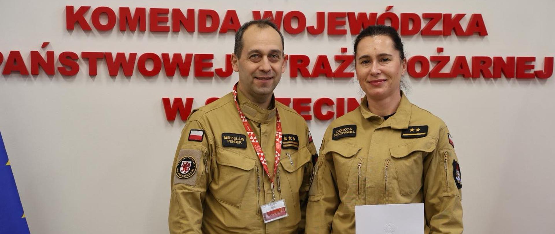 Na zdjęciu dwie osoby w mundurach służbowych stoją w pomieszczeniu urzędowym. Jedna z nich trzyma dokument. W tle widoczny jest napis z nazwą instytucji oraz fragmenty flag Unii Europejskiej i Polski.