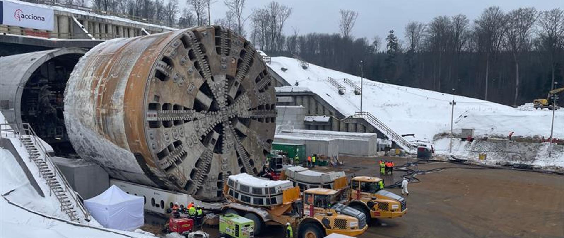 Duża tarcza TBM do drążenia tuneli ustawiona na placu budowy przy wylocie tunelu. Wokół znajdują się maszyny budowlane, pracownicy w odzieży ochronnej oraz zaśnieżony teren.