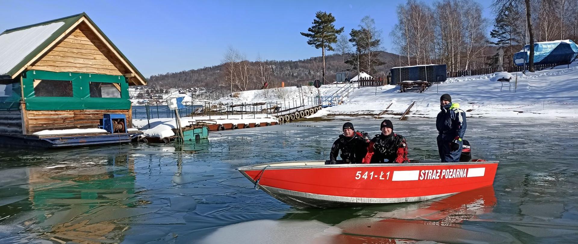 Na zdjęciu trzech strażaków nurków płynących łodzią motorową, dwóch ubranych w sprzęt nurkowy, w tle przystań bazy WOPR w Polańczyku