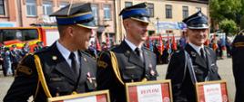 Zdjęcie grupy strażaków w mundurach galowych. W pierwszym planie strażacy trzymający dyplomy w złotych ramach. 