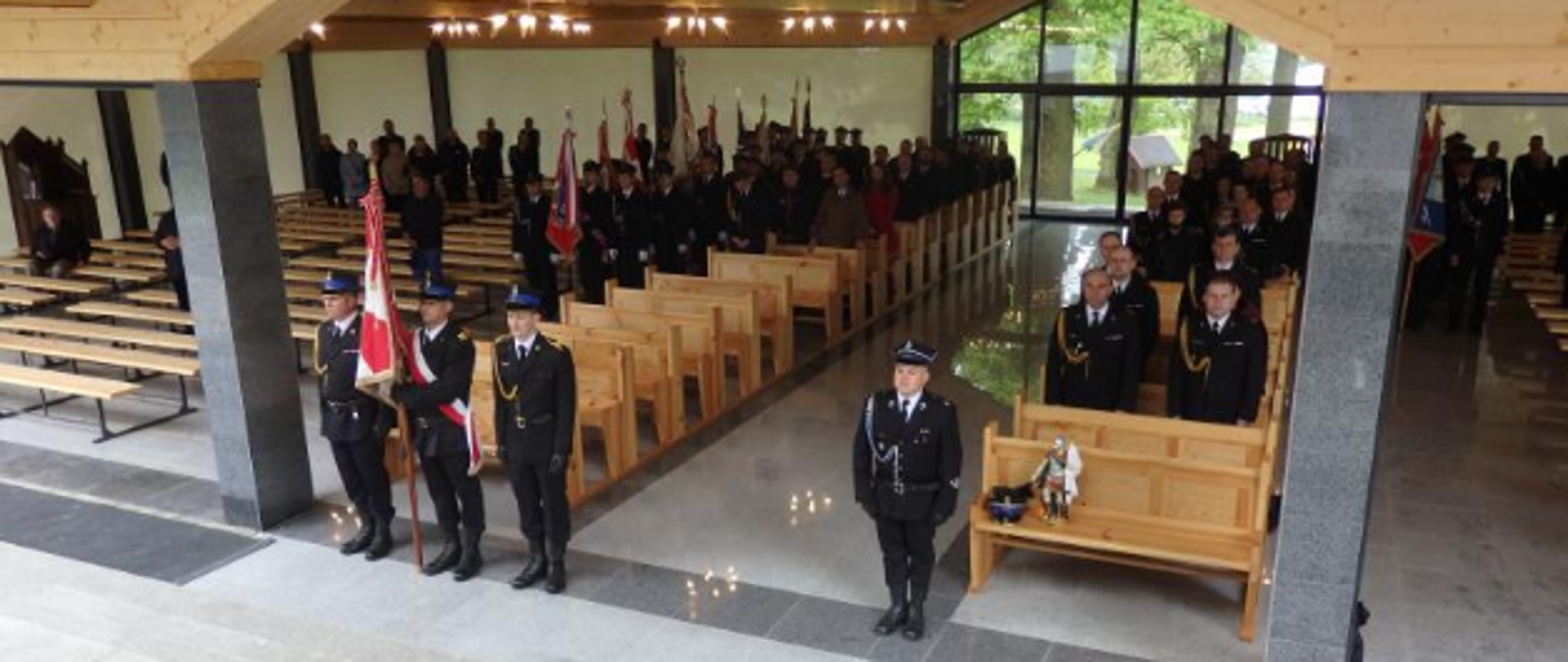 Zdjecie przedstawia osoby znajdujące się w pozycji stojącej w ławkach w zadaszonym pomieszczeniu. W tle widać poczty sztandarowe.