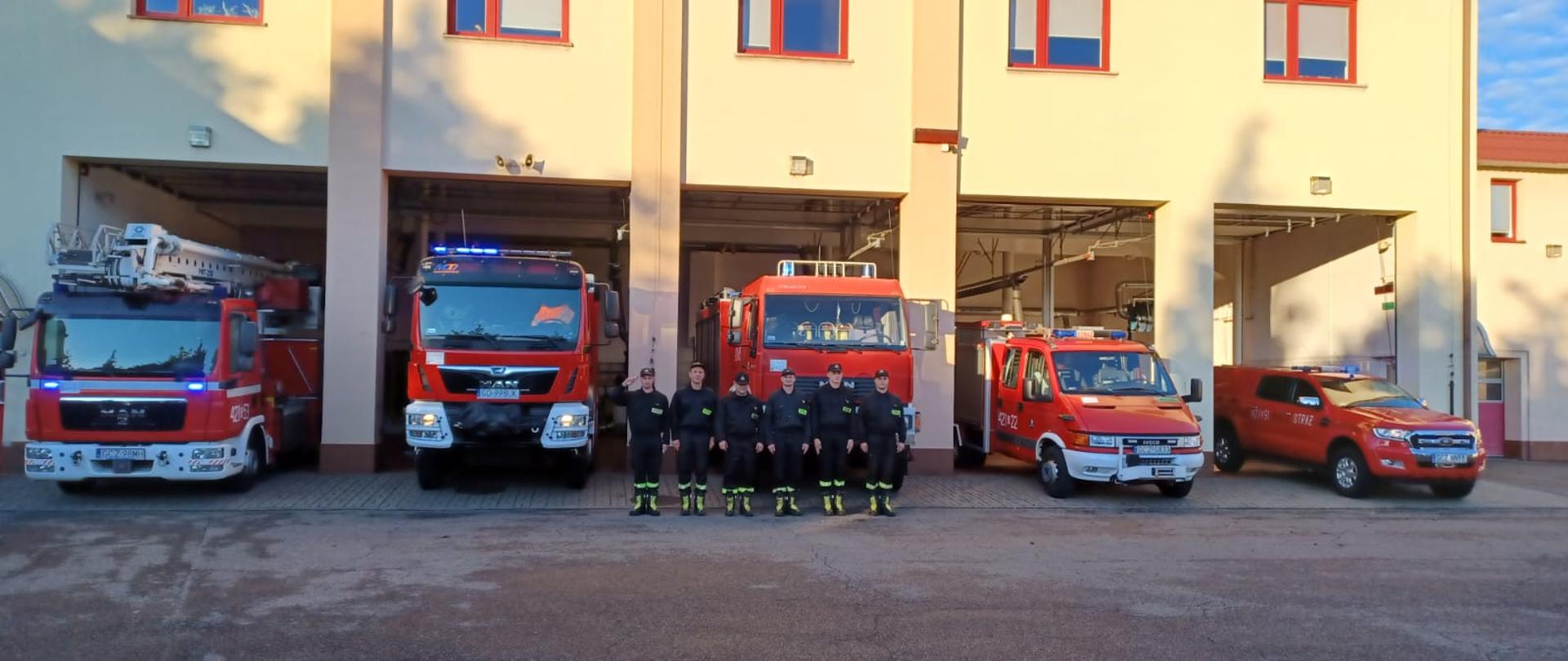 Przed garażem stoją pojazdy strażackie z włączonymi sygnałami dźwiękowymi i świetlnymi. Przed samochodami stoi sześciu strażaków, jeden salutuje.
