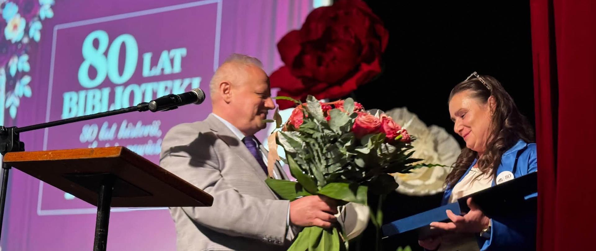 Mężczyzna wręcza kwiaty kobiecie, oboje stoją na scenie, w tle wyświetlany napis 80-lecie Biblioteki 