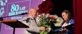 Mężczyzna wręcza kwiaty kobiecie, oboje stoją na scenie, w tle wyświetlany napis 80-lecie Biblioteki 