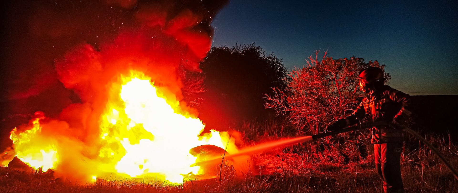 W centrum fotografii znajdują się opony, który intensywnie się spalają. Widoczny jest duży ogień i dym. Po prawej stronie strażak podaje prąd piany na palące się opony. Zdjęcie wykonano przy zmierzchu.
