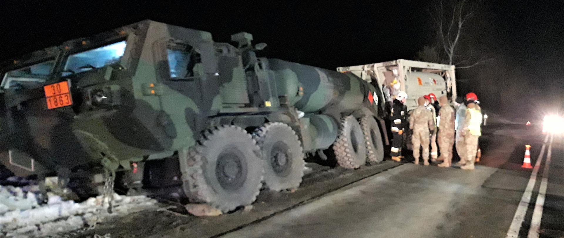 Na zdjęciu widzimy samochód wojskowy-cysternę, który stoi na grząskim poboczu. Widać głębokie koleiny i mocno pogrążone w błocie koła unieruchomionego pojazdu. Za samochodem widać białą przyczepę-cysternę i grupę stojących żołnierzy wraz ze strażakami.