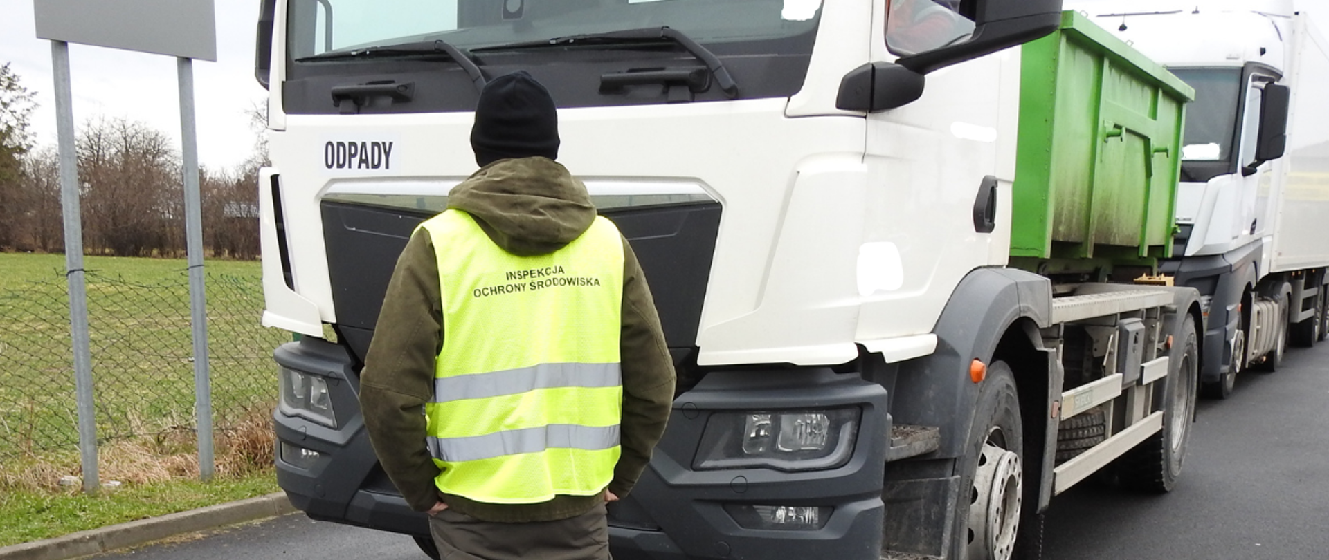 Na pierwszym planie znajduje się osoba obrócona plecami do zdjęcia, ubrana w ciemne spodnie, biało-niebieskie buty sportowe oraz kurtkę z kapturem w ciemnym kolorze, na to założoną ma odblaskową kamizelkę z napisem "Inspekcja Ochrony Środowiska". Na głowie osoba ma czarną czapkę. Przed osobą stoi zaparkowany samochód ciężarowy z zieloną naczepą. Samochód z przodu ma tabliczkę z napisem "Odpady". Za nim stoi kolejny zaparkowany samochód ciężarowy z białą naczepą, za którym stoją zaparkowane samochody osobowe. W tle zdjęcia po lewej stronie widać znak drogowy, a za nim zielone pole i w oddali drzewa. W tle po prawej stronie zdjęcia widać budynek. 