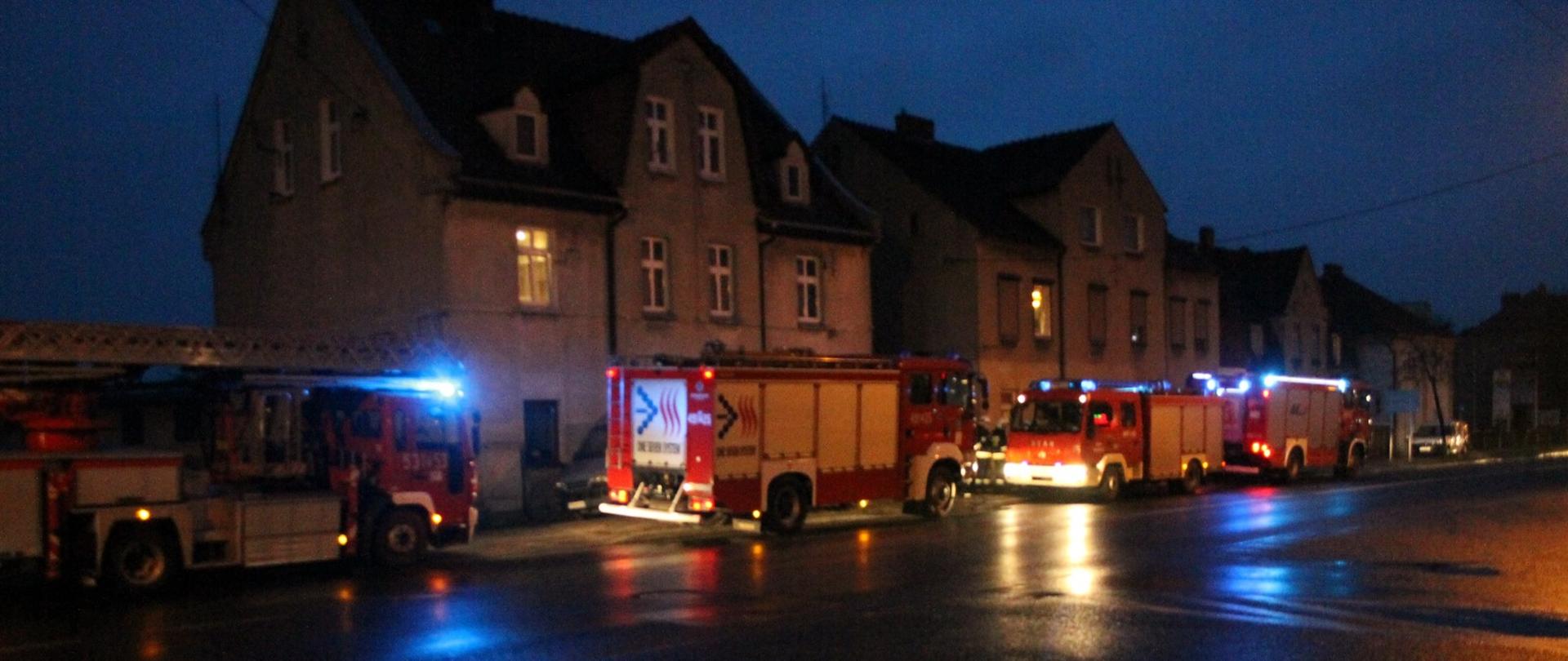 Na drodze przebiegającej przy budynku wielorodzinnym stoją zaparkowane wozy strażackie. Wozy mają włączone sygnały błyskowe. Strażacy prowadzą działania w budynku wielorodzinnym, gdzie stwierdzono występowanie tlenku węgla