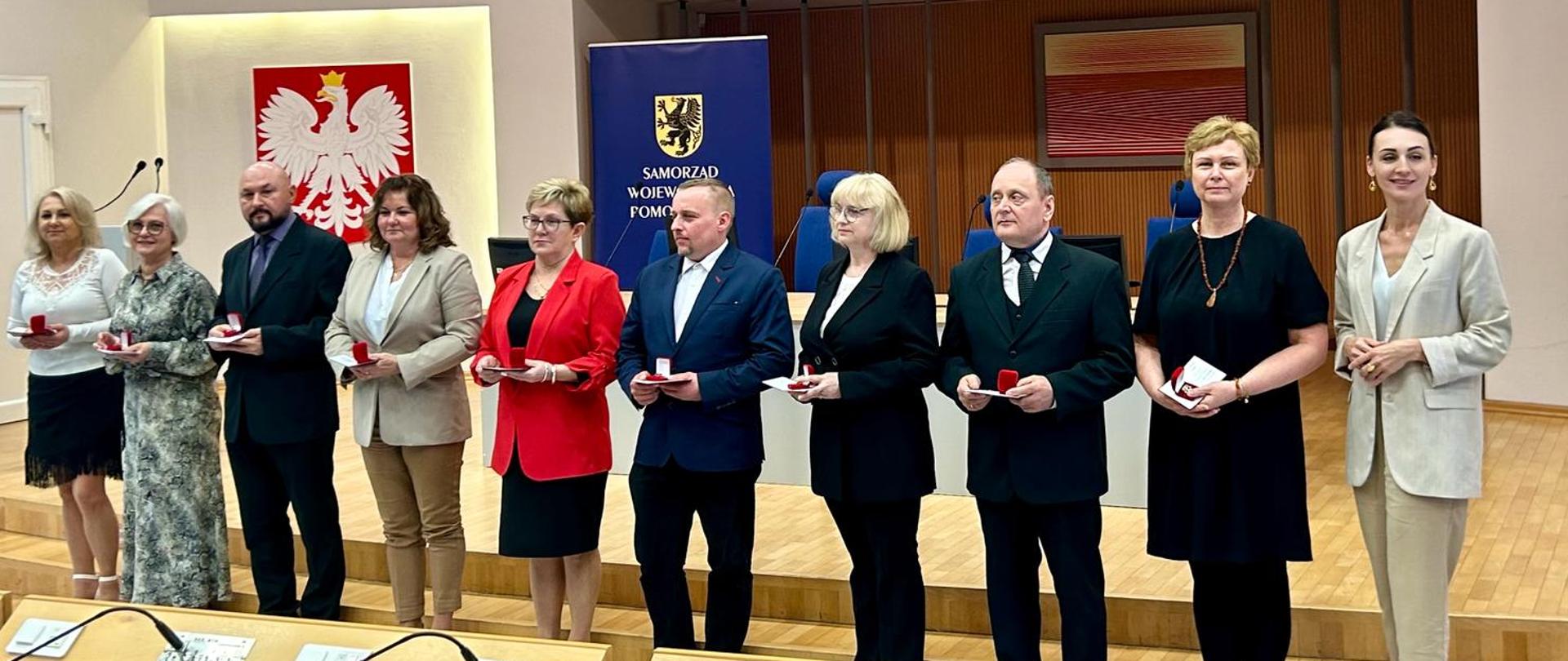 duża sala, w niej wojewoda pomorska i kilka osób (kobiety i mężczyźni) trzymają w dłoniach pudełka z odznakami honorowymi. W tle wizerunek orła