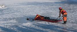 Dwóch strażaków przy pomocy sani lodowych wydobywa z przerębli osobę poszkodowaną (strażaka). W tle aura śnieżna, staw skuty lodem.