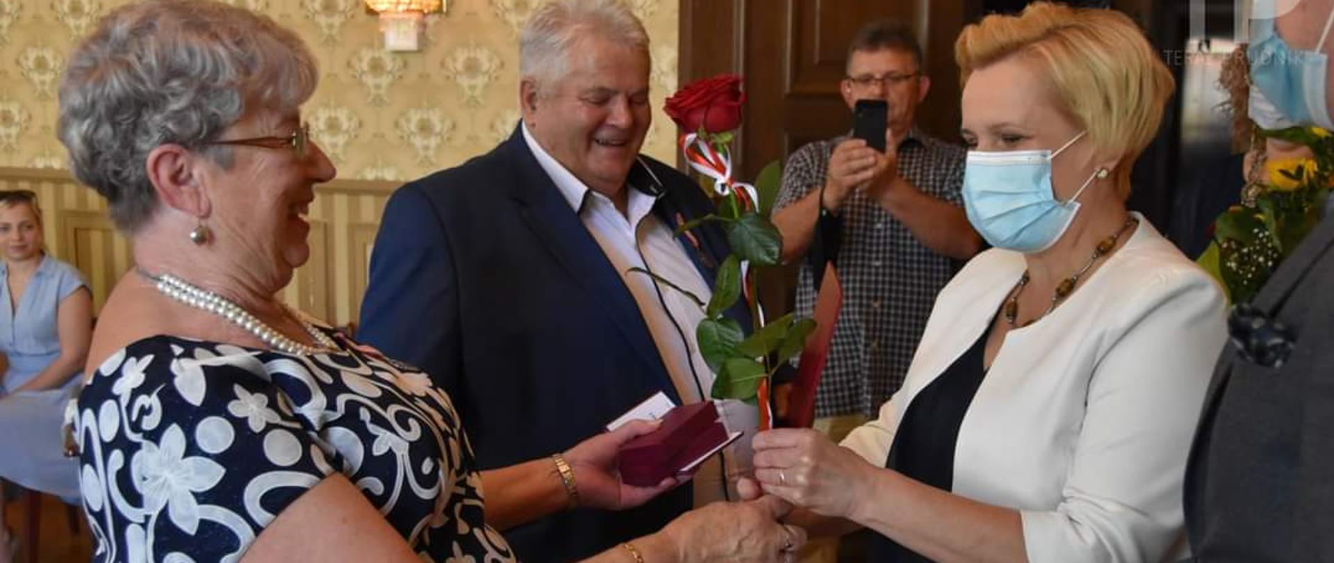 Wicewojewoda opolski wręcza odznaczenie państwowe jednej z wyróżnionych par małżeńskich, które obchodzą Złote Gody. Na zdjęciu po lewej stronie kobieta i mężczyzna w dojrzałym wieku. Na przeciw nich wicewojewoda opolski, która wręcza kobiecie czerwoną różę oraz odznaczenie państwowe nadane przez prezydenta RP. Na skraju kadru po prawej stornie widać zarys mężczyzny. W tle pozostali uczestnicy uroczystości, 