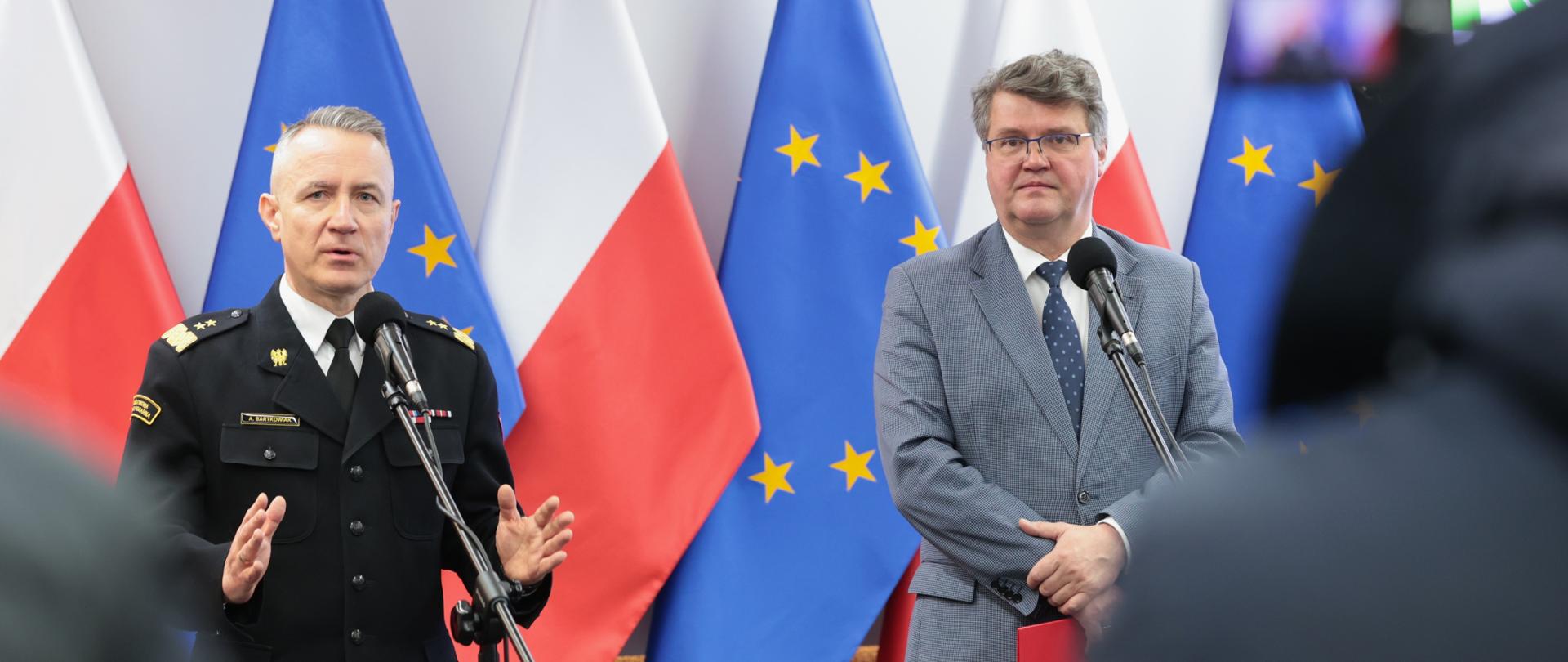 Mężczyzna w strażackim mundurze generalskim oraz drugi w okularach i szarym garniturze stojący przed mikrofonami. Za nim flagi Polski oraz Unii Europejskiej. Na pierwszym planie widoczni dziennikarze biorący udział w konferencji