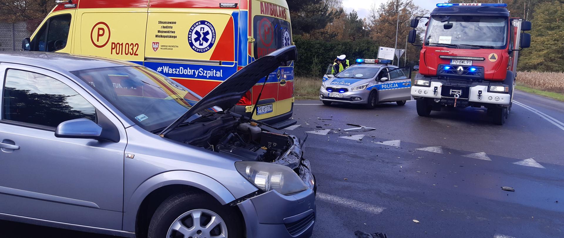 Na zdjęciu widzimy samochód osobowy który brał udział w wypadku drogowym. Samochód koloru srebrnego stoi na środku skrzyżowania, podniesiona przednia maska. Za samochodem osobowym widzimy karetkę pogotowia koloru żółtego. Z prawej strony kadru widzimy samochód strażacki koloru czerwonego, białe zderzaki. Obok samochodu strażackiego stoi policyjny radiowóz.
