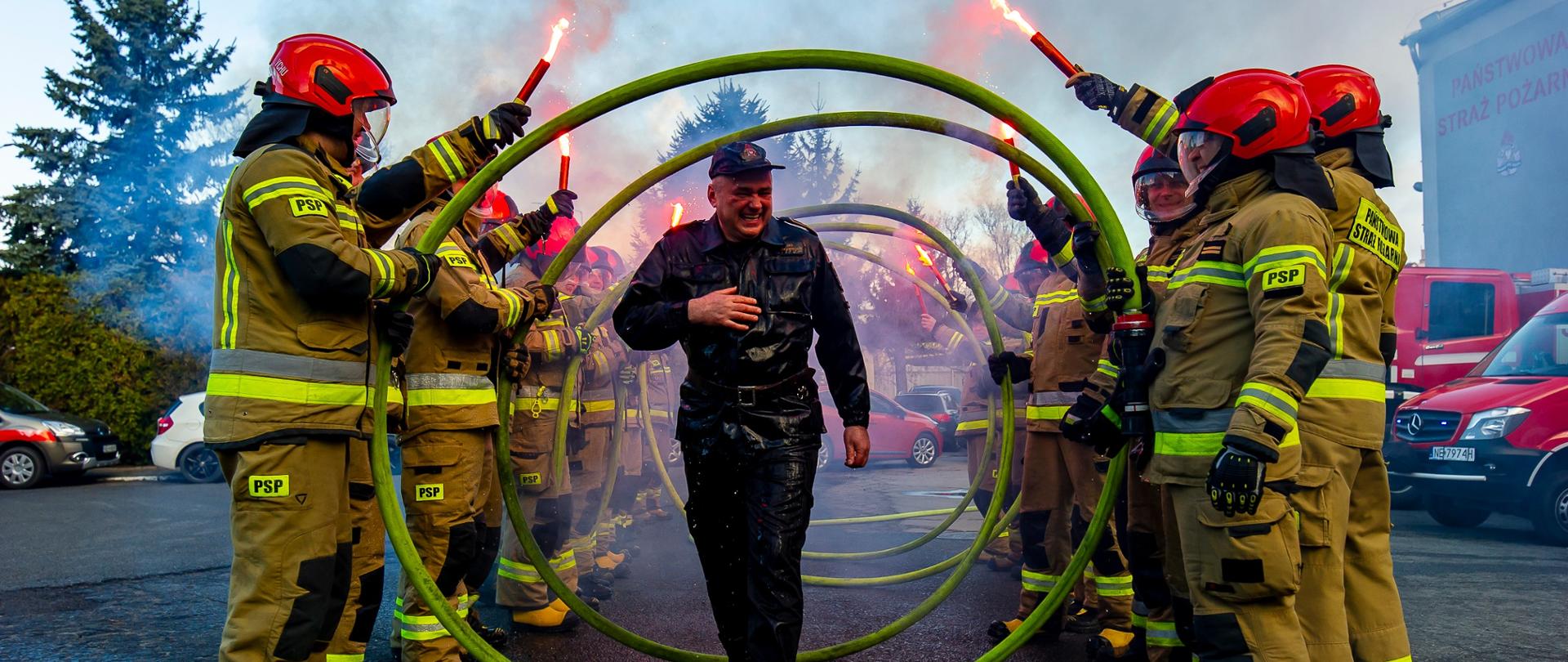 Emerytowany strażak przechodzi przez między dwoma rzędami strażaków. Strażacy w rękach trzymają czerwone flary.