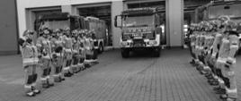 strażacy ustawieni w szeregach na przeciwko siebie stoją na baczność ubrani w stroje specjalne za nimi ustawione są samochody ratowniczo gaśnicze w tle znajduje się budynek Państwowej Straży Pożarnej