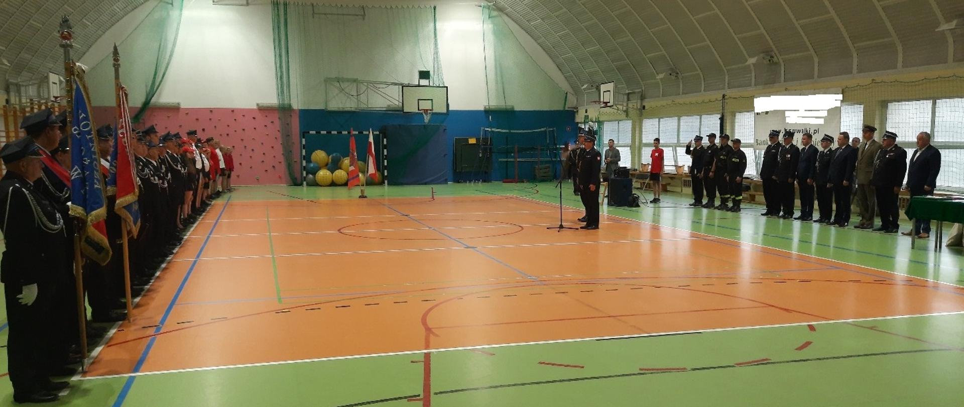 W hali sportowej znajdują się strażacy i inne osoby. Dwóch strażaków jest przy mikrofonie.