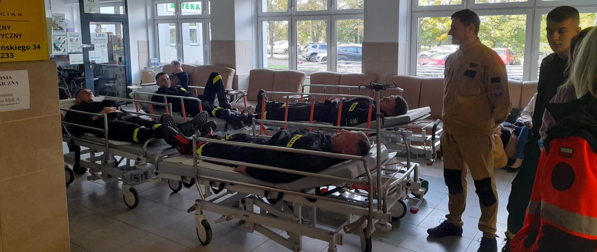 Na zdjęciu znajduję się grupa strażaków biorących udział w pozoracji, którzy leżą na łóżkach w sali szpitalnej podczas ćwiczeń, obok nich znajduje się ratownik medyczny oraz pacjenci.