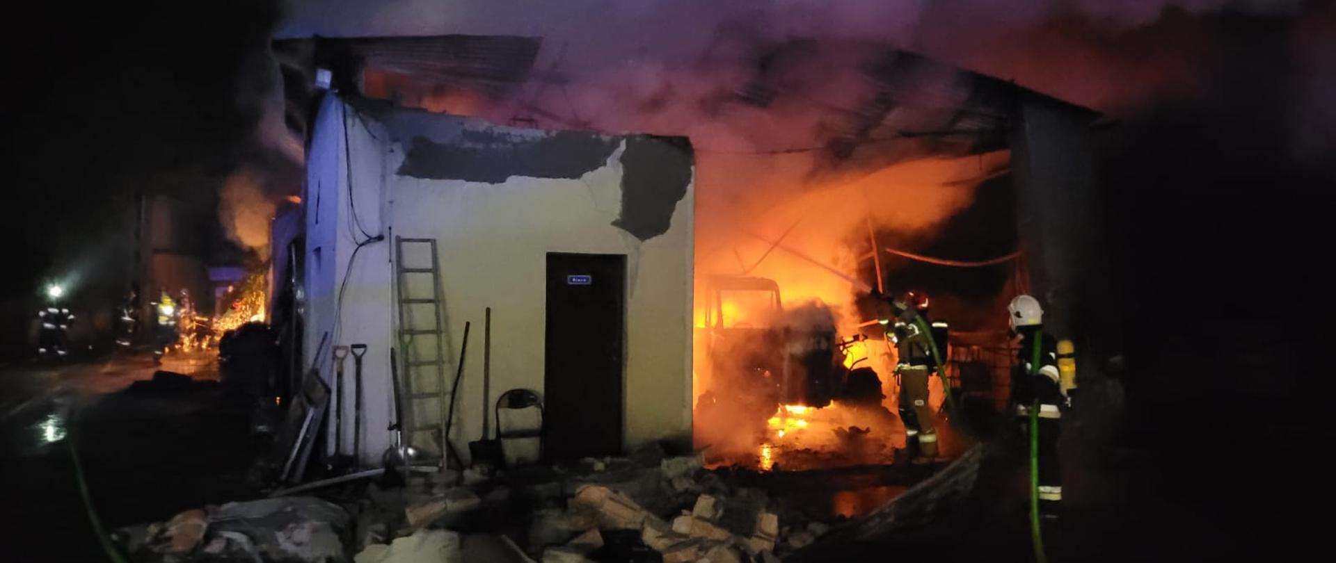 Na zdjęciu widać budynek produkcyjny podczas pożaru, w środku maszyny objęte ogniem, dach budynku zawalony do środka, na pierwszym planie dwóch strażaków (1 OSP i 1 PSP) w ubraniach bojowych i aparatach ochrony układu oddechowego, podających wodę na palący się budynek, w tle widać strażaków w ubraniach specjalnych prowadzących prace rozbiórkowe.