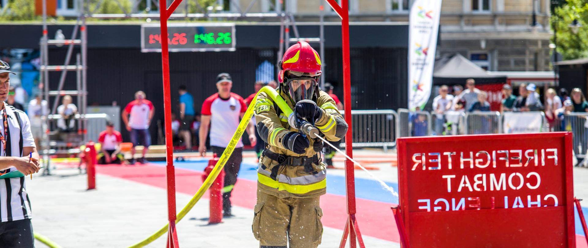 Zdjęcie przedstawia strażaka - uczestnika zawodów w pełnym umundurowaniu specjalnym wraz z aparatem ODO podającego prąd wody na tarczę. Na zdjęciu ujęto także widzów oraz sędziów zawodów.