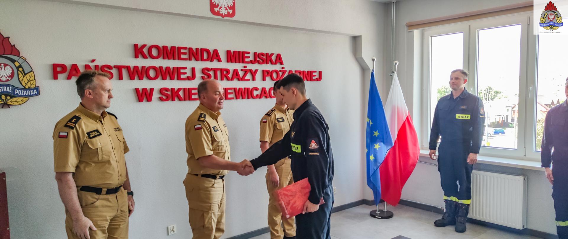 Strażacy w mundurach koloru piaskowego oraz czarnego stoją przy ścianie, jeden odbiera dokument.