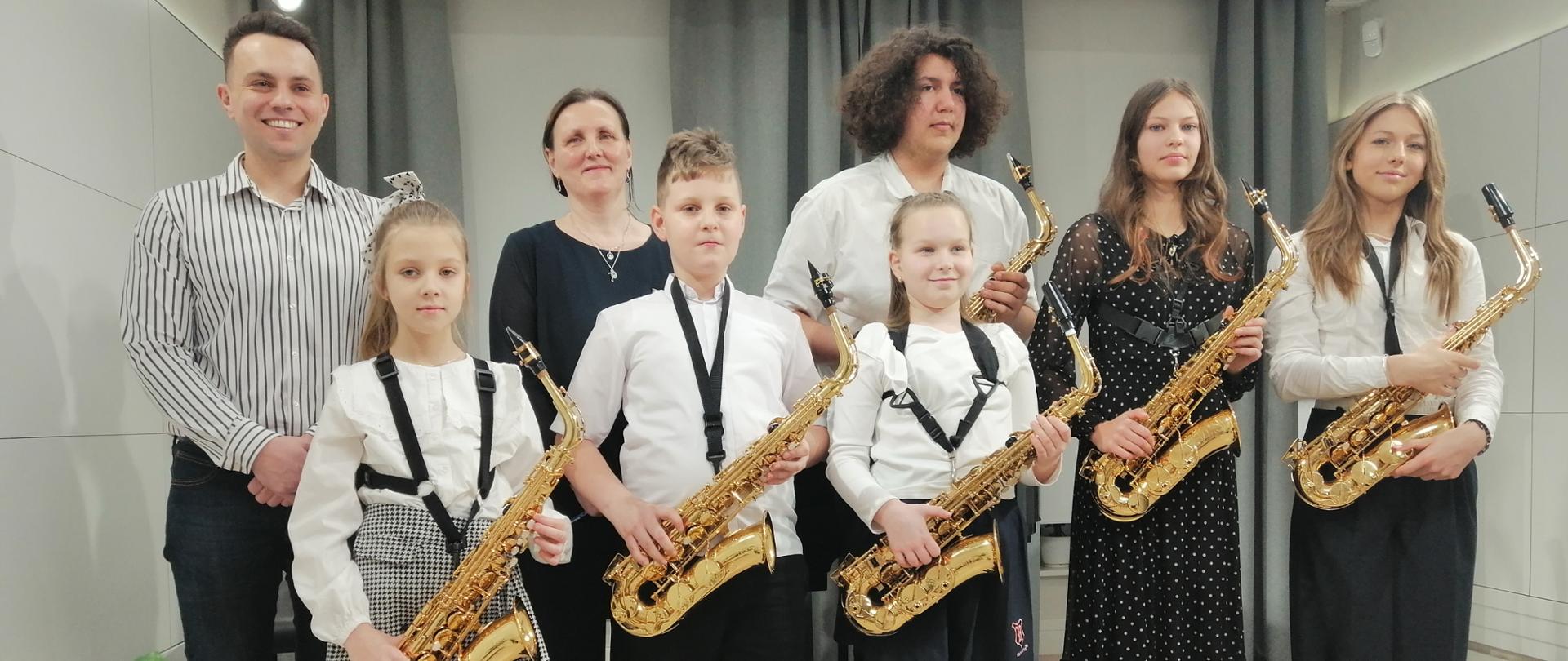 Zdjęcie grupowe uczniów trzymających w rękach saksofony. Uczniowie ubrani są na galowo. Stoją na scenie w dwurzędzie. Z lewej strony stoi nauczyciel, a obok akompaniatorka. 