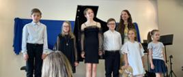 Na zdjęciu widoczna jest grupa dzieci oraz nauczycielka stojący na scenie po zakończonym występie muzycznym. W tle znajduje się fortepian koncertowy. Uczniowie ubrani są elegancko — dziewczynki mają sukienki w jasnych i ciemnych kolorach, a chłopcy białe koszule i ciemne spodnie. Wszyscy stoją obok siebie i patrzą w stronę publiczności. Na pierwszym planie widać fragment widowni. Atmosfera zdjęcia jest uroczysta i radosna, charakterystyczna dla szkolnego koncertu fortepianowego.