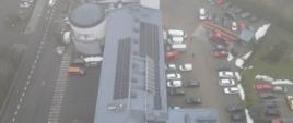 Zdjęcie przedstawia widok z góry na dach obiektu jednostki ratowniczo - gaśniczej numer 1 w Radomiu. Na zdjęciu widać zamontowane panele instalacji fotowoltaicznej. Dookoła obiektu widać zaparkowane pojazdy zarówno osobowe jak i pożarnicze. Zdjęcie wykonane w okresie zimowym.