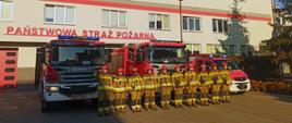 Na zdjęciu widocznych jest 10 strażaków w piaskowych ubraniach specjalnych i czerwonych hełmach. Za strażakami stoją trzy samochody strażackie z włączonymi sygnałami błyskowymi. Na drugim planie budynek sochaczewskiej straży pożarnej.