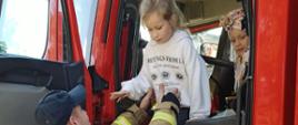 Na zdjęciu widać troje dzieci w wieku przedszkolnym i jednego strażaka. Dwoje z nich znajduje się w kabinie wozu strażackiego. Strażak pomaga wyjść z samochodu jednej dziewczynce. O wóz strażacki oparty jest szyld Kampanii prewencyjnej "Kręci mnie bezpieczeństwo przez cały rok szkolny".
