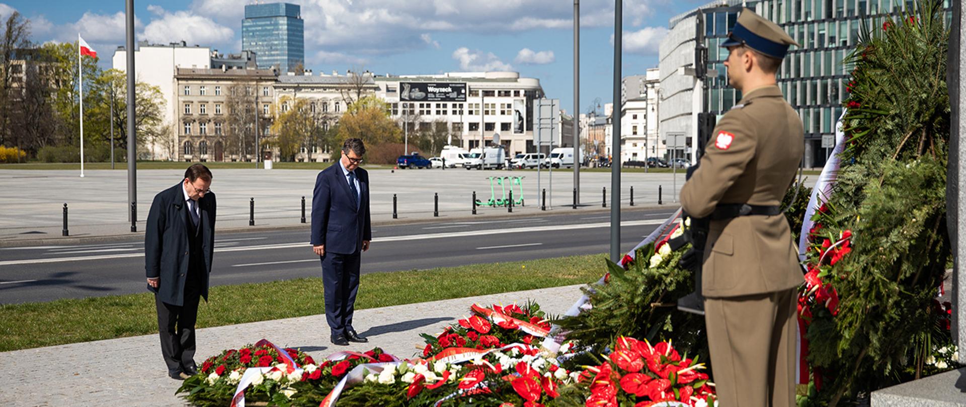 Na zdjęciu widać stojących z pochylonymi głowami Mariusza Kamińskiego ministra SWiA i Macieja Wąsika wiceministra SWiA przed pomnikiem Lecha Kaczyńskiego prezydenta RP w Warszawie. Widać również jednego z żołnierzy pełniących wartę przy pomniku i wiele złożonych wieńców