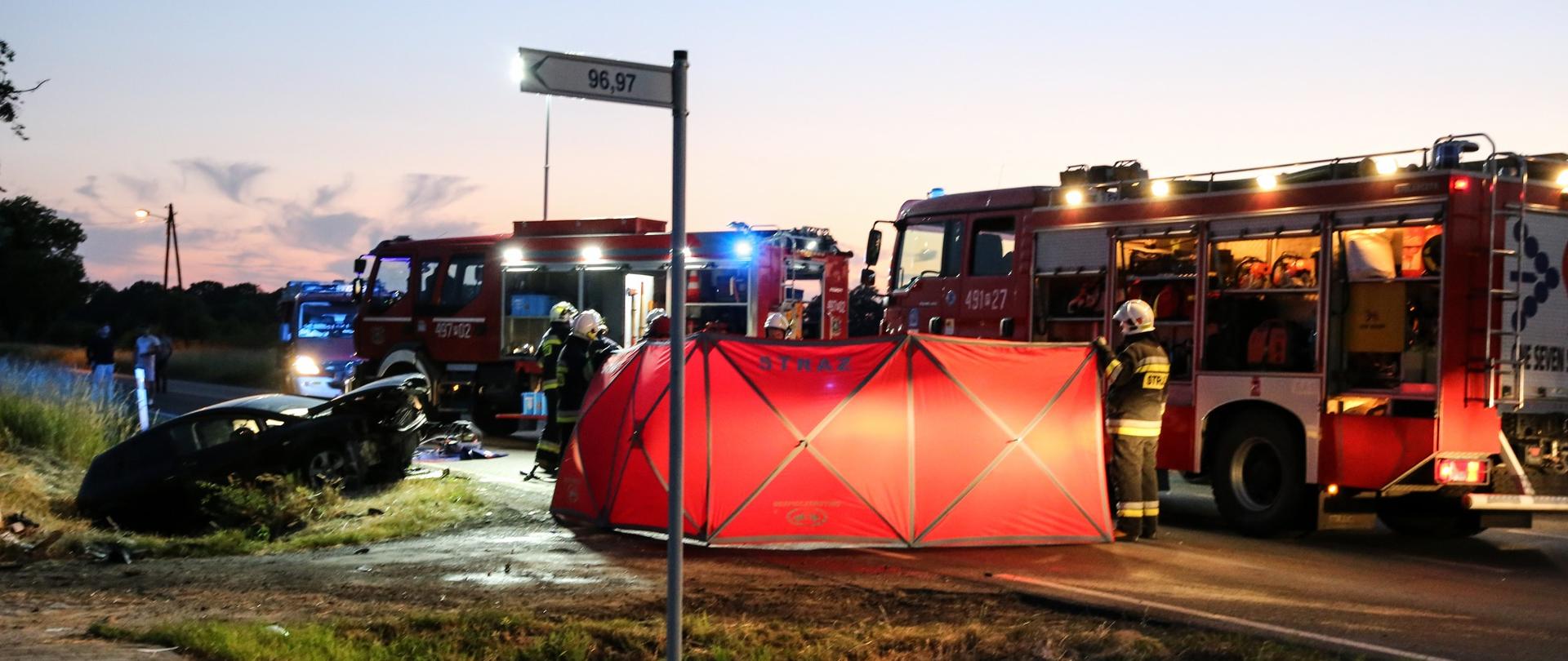 Zdjęcie przedstawia samochody pożarnicze stojące na drodze, parawan ratowniczy koloru czerwonego oraz rozbity pojazd biorący udziała w zdarzeniu. Przy wozach strażackich stoją strażacy w umundurowaniu bojowym.