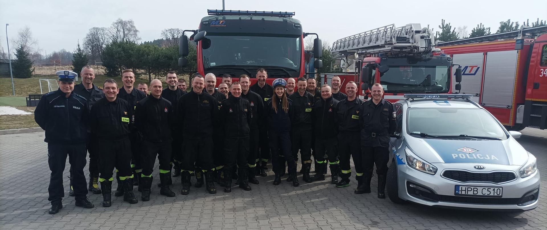 Na zdjęciu widać strażaków razem z policjantami. Funkcjonariusze stoą na placu przed jednostką. Obok funkcjonariuszy stoją pojazdy policyjne oraz strażackie. 