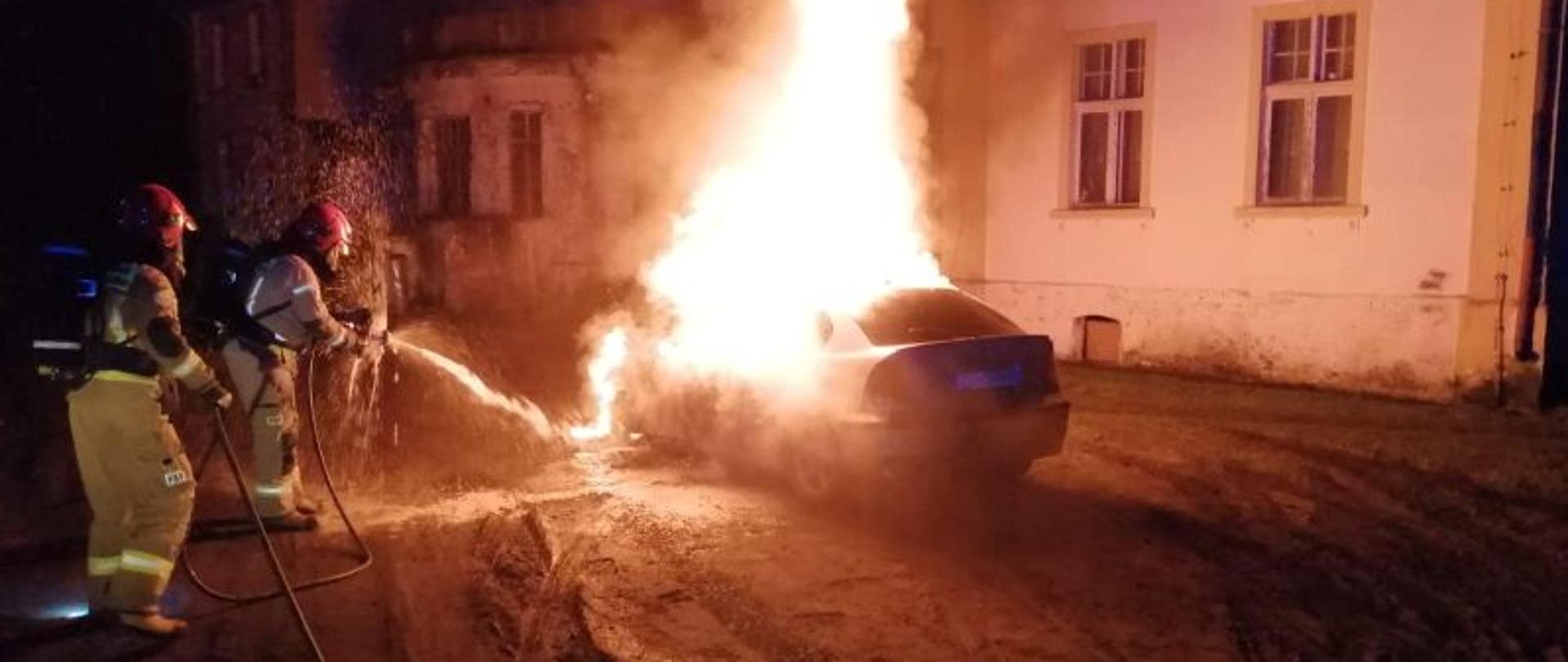 Zdjęcie wykonane w nocy. Centralny punkt zdjęcia stanowi palące się auto. Auto całkowicie objęte ogniem. Ogień bardzo duży. Żółty, czerwony, pomarańczowy. Strażacy przy aucie podają prąd piany na palący się obiekt.