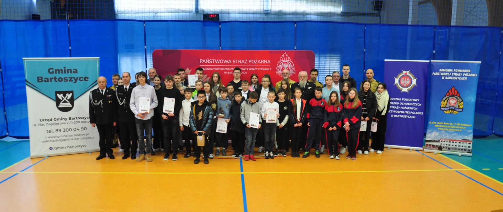 Wszyscy uczestnicy konkursu i organizatorzy na wspólnym zdjęciu na tle banerów Państwowej Straży Pożarnej w Bartoszycach, Gminy Bartoszyce i Oddziału Powiatowego Związku Ochotniczych Straży Pożarnych