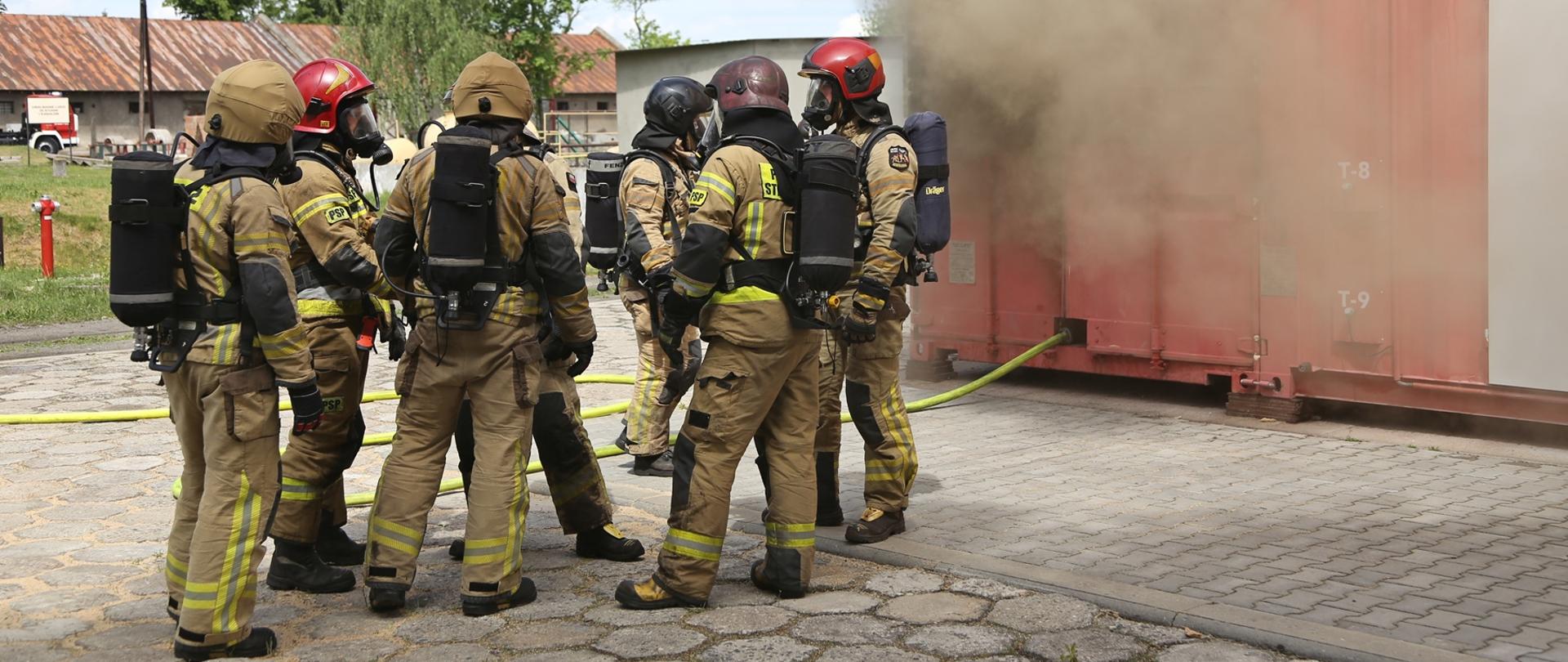 Po lewej strażacy w umundurowaniu gaśniczym, z założonymi butlami (siedem osób), po prawej kontener gaśniczy z którego wydobywa się dym