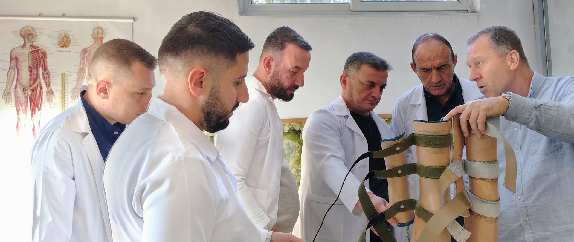 Sześć osób stoi wokół modelu dwóch nóg umieszczonego na stole. Jedna osoba wskazuje elementy konstrukcji z paskami i zapięciami, a pozostałe obserwują prezentację. Cztery osoby mają białe fartuchy. Na ścianie w tle znajduje się plansza przedstawiająca budowę anatomiczną człowieka.