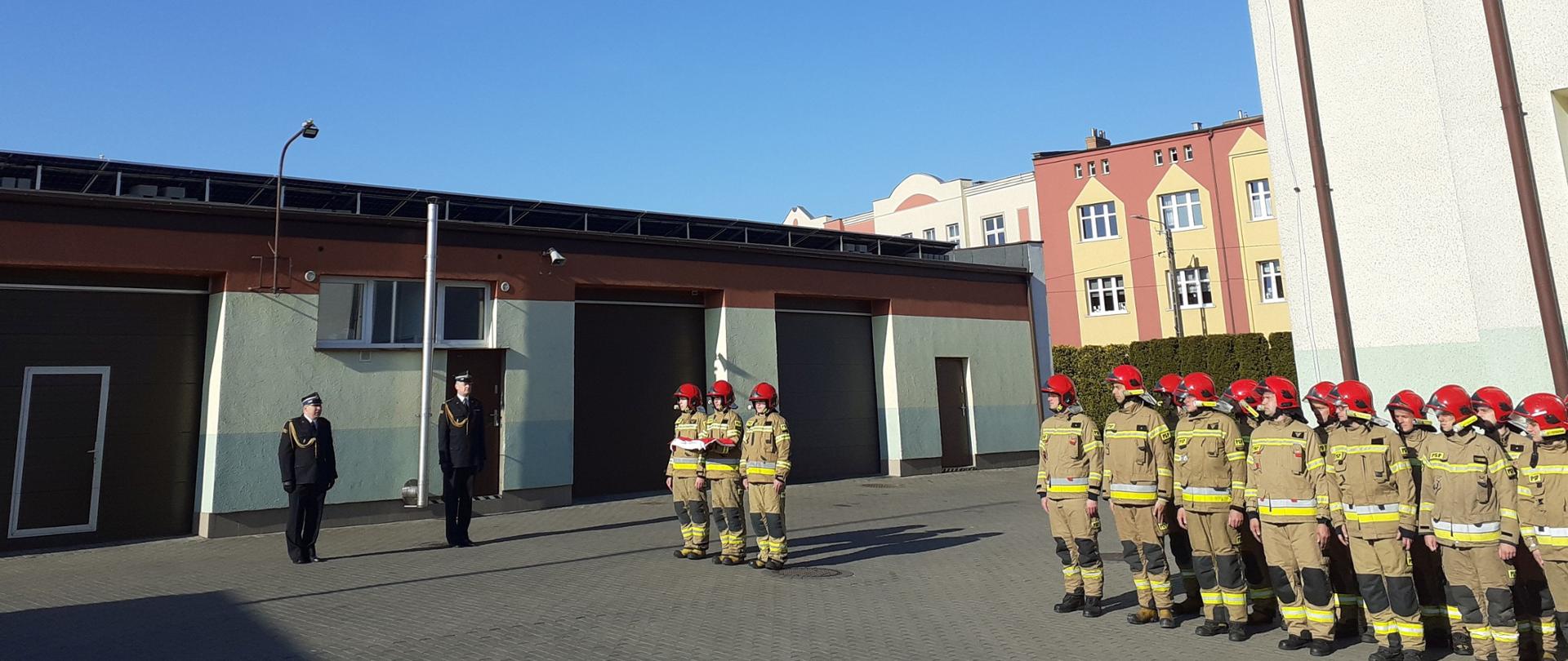 Zdjęcie przedstawia uroczystość podniesienia flagi państwowej, strażacy ustawieni w szeregu, poczet flagowy z flagą zmierza do masztu, w tle Komendant i dowódca.