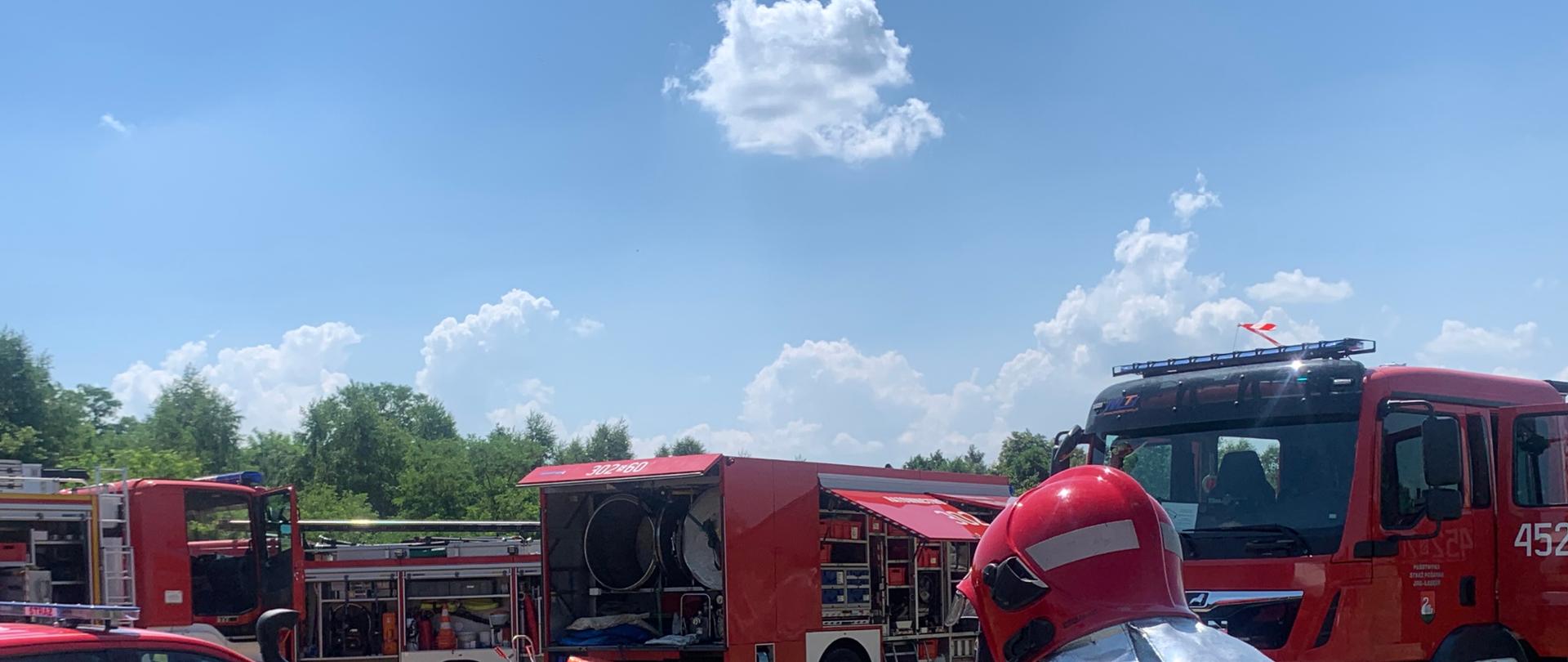 Zdjęcie przedstawia Kierującego Działaniem Ratowniczym (ćwiczeniami) na tle pojazdów pożarniczych