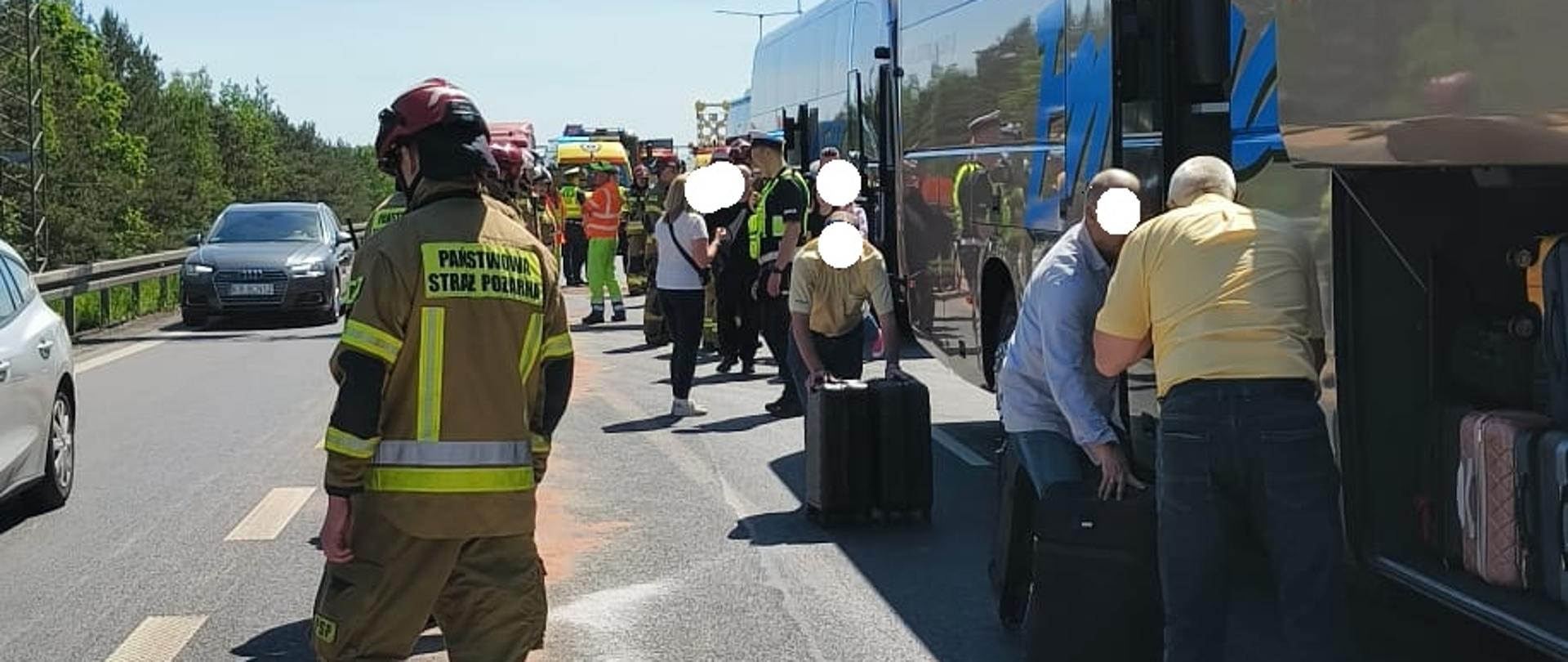 Zdjęcie przedstawia akcję ratunkową na autostradzie, gdzie obok strażaka i pasa wysypanego sorbentu pasażerowie wyciągają i zabierają swoje walizki z luków bagażowych stojących autokarów.
