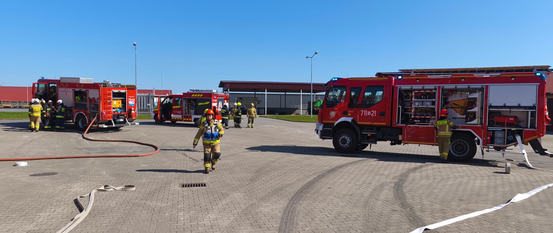  placu manewrowym widać kilka zastępów straży pożarnej oraz strażaków w pełnym umundurowaniu bojowym, którzy rozwijają węże gaśnicze. Scena przedstawia ćwiczenia służb ratowniczych w słoneczny, bezchmurny dzień.
