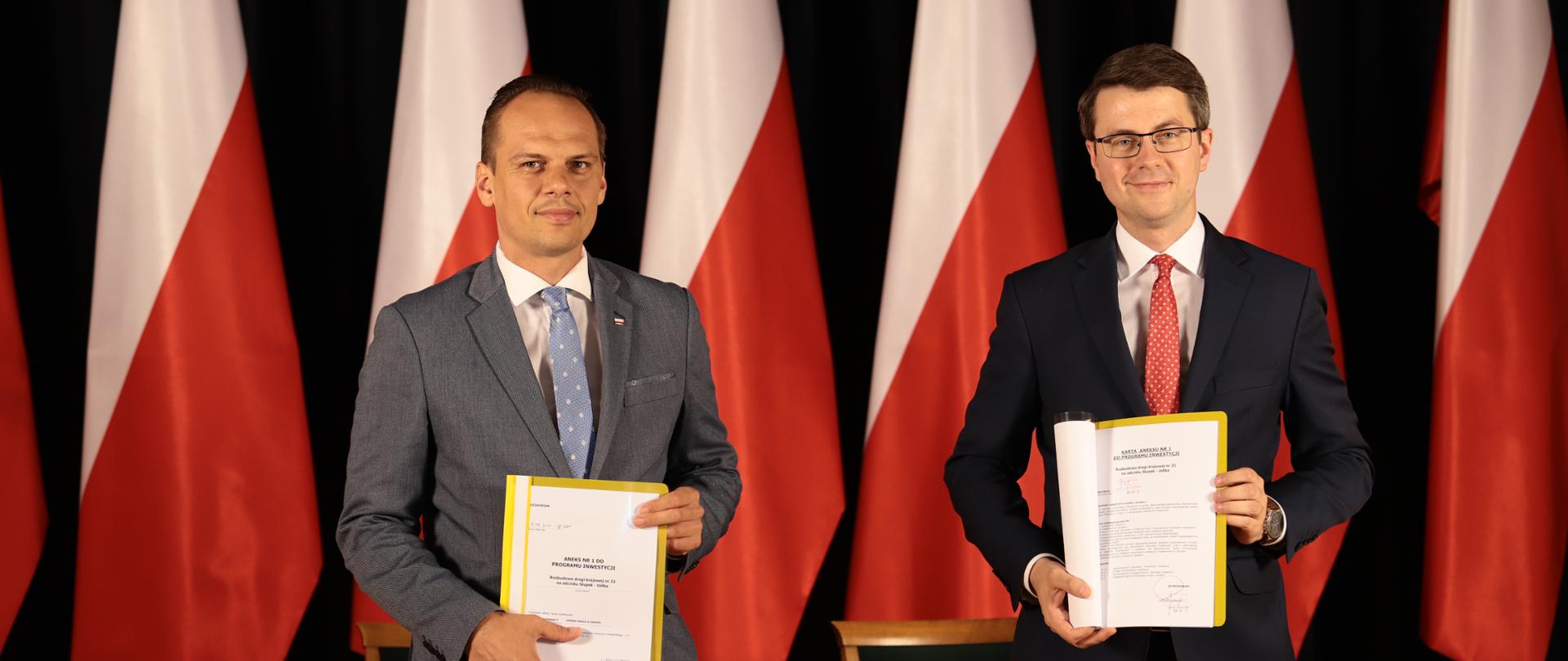 Na zdjęciu widać dwóch mężczyzn ubranych w garnitury. Panowie są uśmiechnięci, każdy z nich trzyma dokument w ręku. W ich tle widać brązowy stół oraz sześć biało-czerwonych flag.