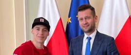Minister Kamil Bortniczuk a obok żużlowiec Bartłomiej Kowalski. Minister ubrany jest w granatowy garnitur w kratkę, a żużlowiec w czerwoną bluzę reprezentacyjną z orzełkiem na piersi. 